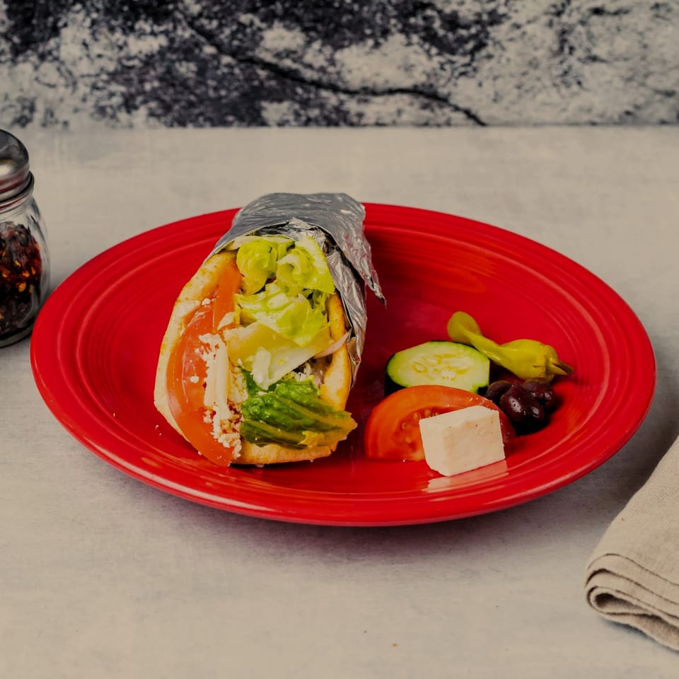 A gyro wrapped in foil on a red plate with feta, olives, cucumbers, tomatoes, and peppers.