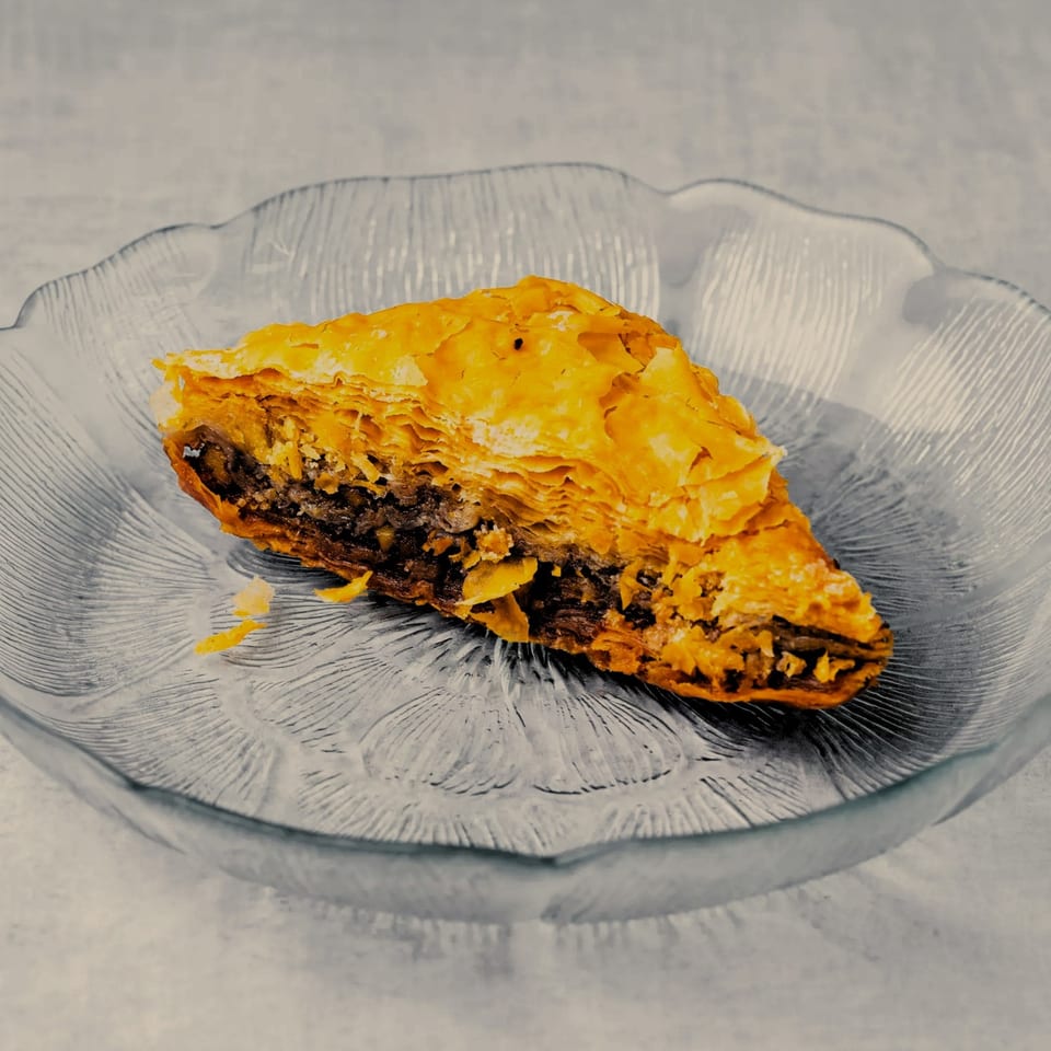 A triangular slice of baklava with flaky golden crust and dark filling sits on a clear glass plate.