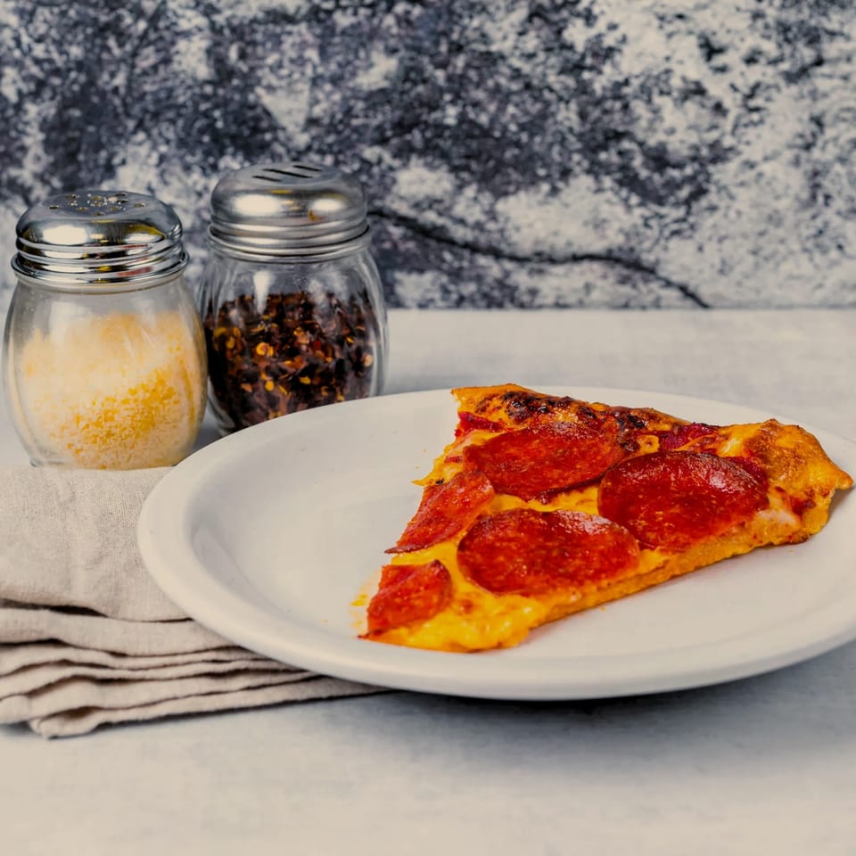 A slice of pepperoni pizza on a white plate with parmesan and red pepper flakes.