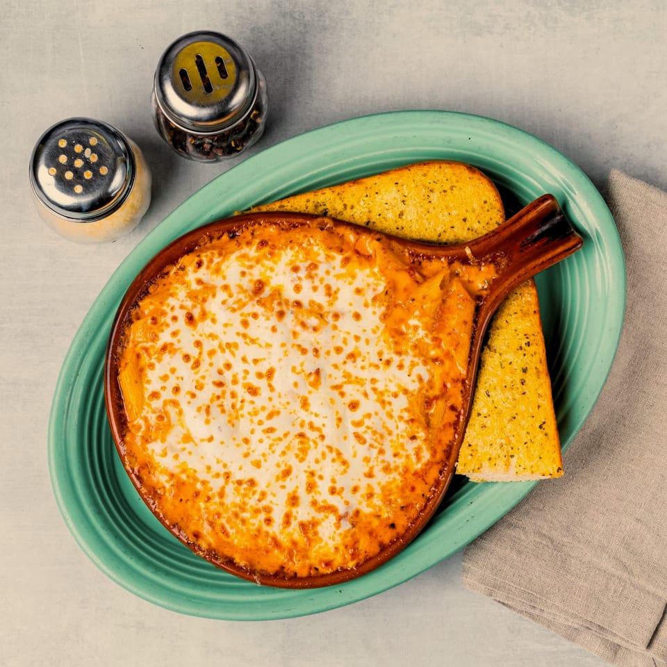 Baked pasta with melted cheese in a brown dish, garlic bread, and shakers on a teal plate.